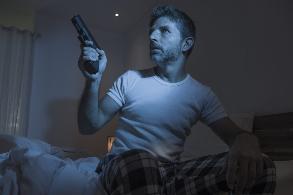 night edgy portrait of young stressed and paranoid American man sitting on bed unable to sleep holding gun looking around scared feeling threatened suffering paranoia  in self protection concept