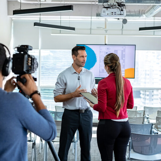 Happy businessman during corporate interview with female journalist. Manager answering question in office. Young woman at work as reporter with business man and cameraman shooting video for broadcast